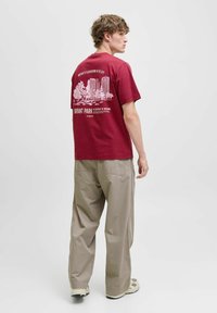 Bordeaux bomulds T-shirt med grafisk print af bygninger og træer på ryggen. Kombineret med beige vidde bukser og sneakers.