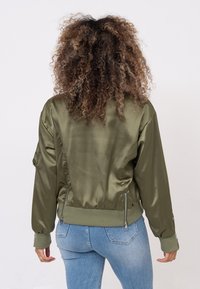 Blouson aviateur en satin vert olive avec poignets et ourlet côtelés, doté de fermetures éclair latérales et d'une coupe décontractée. Texture lisse et design structuré.