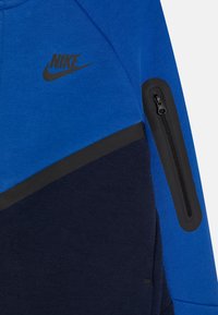 Blauer Sweatshirt mit einem schwarzen Nike-Logo, strukturiertem Stoff und einer Reißverschlusstasche am linken Ärmel mit einem schwarzen Akzentband.