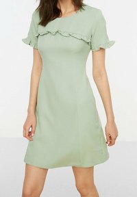 Kvinna iklädd en kortärmad, ljusgrön klänning med volanger över bröstet och ärmsluten, stående mot en enkel bakgrund.