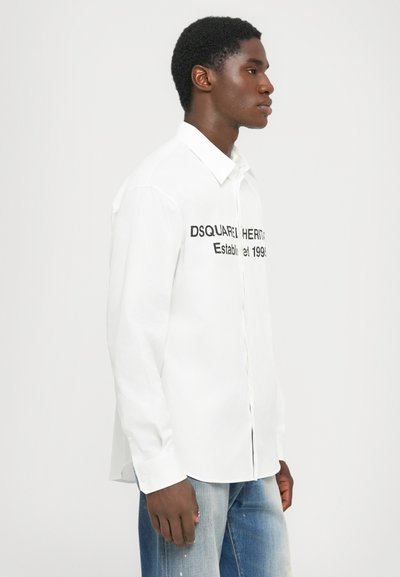 Balta pogājama krekla dizains ar melniem uzrakstiem "DSQUARED HERITAGE Established 1999" priekšpusē, pieskaņots piegriezums un garas piedurknes.