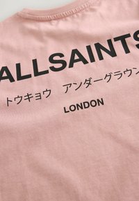 Rožinė medvilninė marškinė su ryškiu juodu tekstu "ALLSAINTS" ir "LONDON" ant nugaros, su papildomu tekstu japonų kalba.