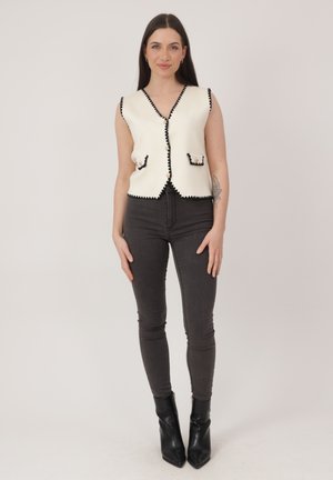 Vrouw staand, draagt een mouwloos crèmekleurig vest met zwarte bies, donkere skinny jeans, zwarte enkellaarsjes en een tatoeage op de linker onderarm.