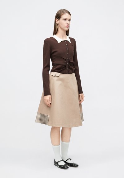 Καφέ ριμπ πλεκτή ζακέτα με λευκό γιακά, συνδυασμένη με μπεζ φούστα σε γραμμή Α με καρό τελείωμα, μαύρα παπούτσια και γκρι κάλτσες.