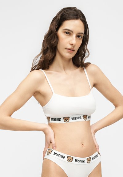 Γυναίκα που φοράει λευκό bralette Moschino και ταιριαστό εσώρουχο με ταινίες με το λογότυπο αρκούδας, στέκεται με τα χέρια στη μέση μπροστά σε απλό φόντο.