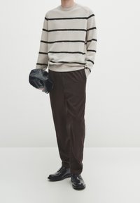 Gestreifter beiger Strickpullover mit schwarzen Streifen, braune weit geschnittene Hose und schwarze Schuhe, hält eine schwarze weiche Tasche. Minimalistisches Design, glatte Textur.