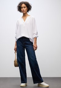 Blusa bianca, ampia e fluida, con scollatura a V e maniche arrotolate, abbinata a jeans larghi blu scuro e sneakers bianche. Porta una borsa beige.