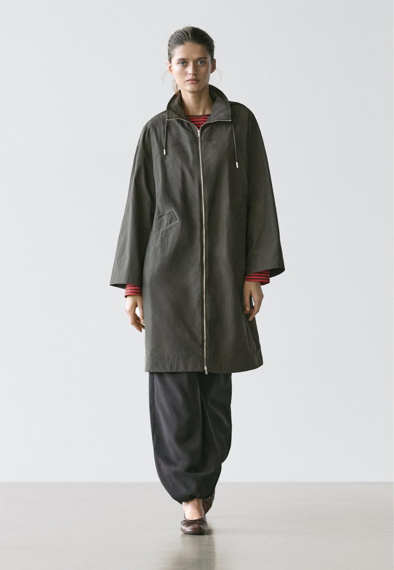 Woman wearing oversized dark green zip-up coat over red striped top, loose black pants, and brown ballet flats walking forward.