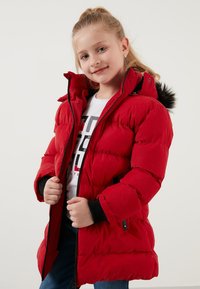 Rød polstret parka-jakke med lynlåslukning, stor hætte med faux fur-kant, ribbede manchetter og sidelommer. Modellen bærer en hvid t-shirt.