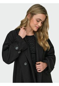 Juodas vilnonis paltas su per dideliu siluetu, dideliais mygtukais ir plačiomis rankovėmis. Dėvimas ant juodos palaidinės su baltais taškeliais. Lygus paviršius.