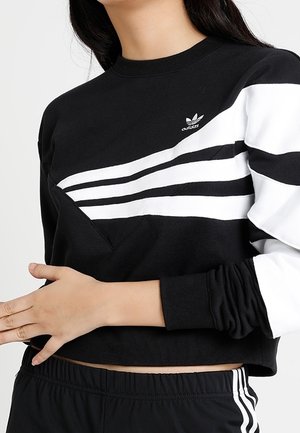 Vrouw draagt een zwarte Adidas cropped sweatshirt met witte strepen en logo, gecombineerd met zwarte Adidas broek met witte strepen.