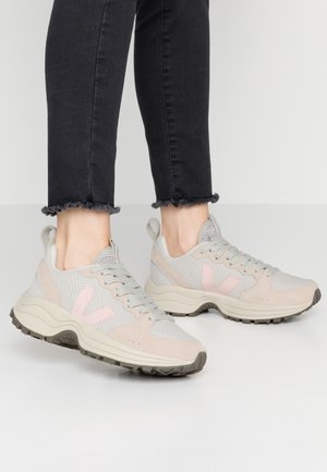 Persoon wearing lichtgrijze en beige mesh sneakers met een roze "V" logo en zwart versleten jeans met rafelige zoom, staand op een witte vloer.