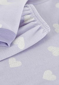 Tessuto lavanda morbido con motivi a cuore bianchi, che mostra il polsino elastico e il dettaglio della manica piegata su un capo d'abbigliamento accogliente.