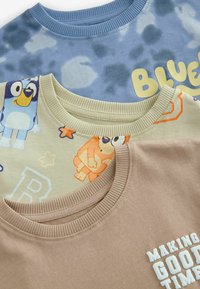 Three t-shirts: a blue tie-dye design, a cream shirt with cartoon characters, and a brown shirt with white text. All have ribbed necklines.
