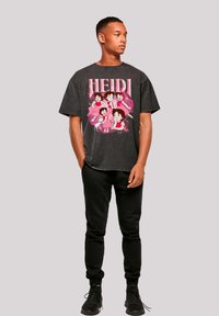Black graphic t-shirt featuring cartoon characters from "Heidi" in pink, paired with black joggers and black athletic shoes, standing on a gray background.
