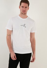 White cotton t-shirt with short sleeves, featuring a black graphic text design on the front. Straight cut hem, worn with black pants.