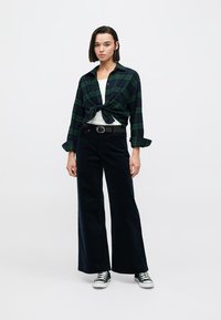 Zeleno-tmavomodrá károvaná košeľa zviazaná v páse, pod ňou biele tričko, doplnené o široké čierne nohavice a čierne tenisky Converse.