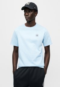 Ανοιχτό μπλε βαμβακερό t-shirt με στρογγυλή λαιμόκοψη και κοντά μανίκια, με κυκλικό λογότυπο στο αριστερό στήθος. Φορεμένο με σκούρο αθ λητικό παντελόνι και ριγέ καπέλο.