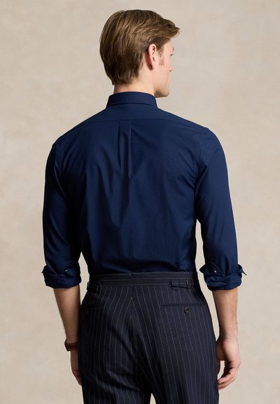 Mężczyzna z podwiniętymi rękawami, ubrany w granatową koszulę i ciemne spodnie w cienkie prążki, zwrócony tyłem na jednolitym beżowym tle.