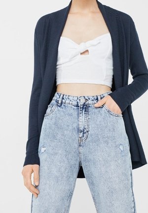 Femme portant un cardigan bleu marine sur un crop top blanc noué à l'avant, associé à un jean taille haute délavé clair, une main dans la poche.