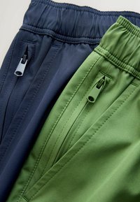 Duonos tamsiai mėlynos ir alyvuogių žalios spalvų reguliuojamo juosmens kelnės. Kiekvienos kelnės turi užtrauktuko kišenę ir glotnią, lengvą audinio tekstūrą.