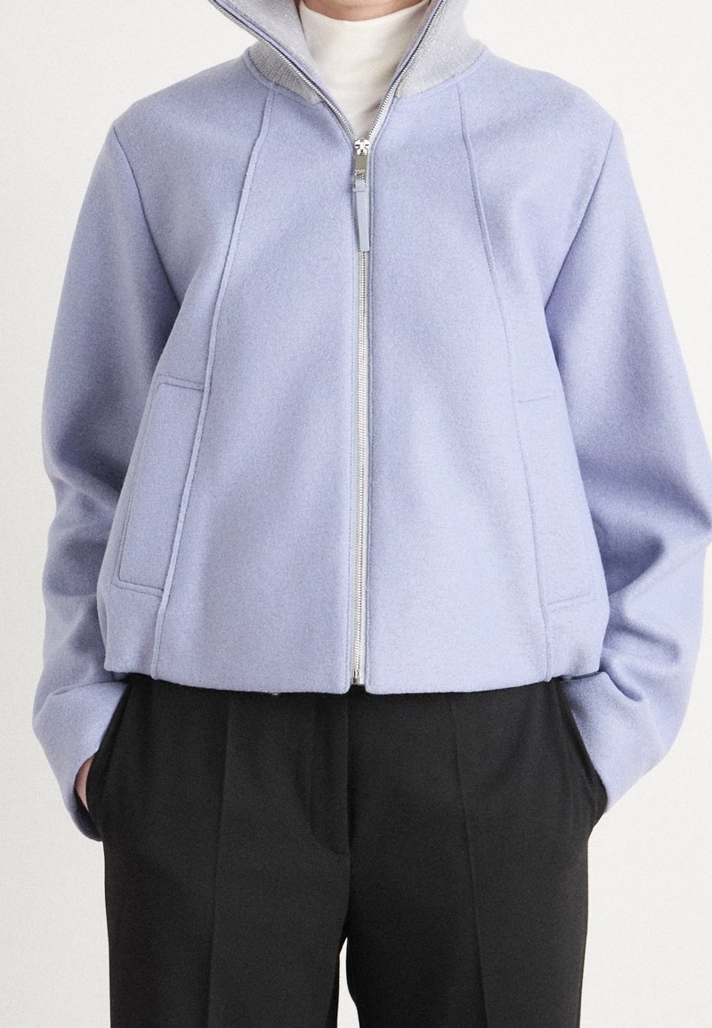 Personne portant une veste zippée violet clair par-dessus un haut blanc et un pantalon noir, les mains dans les poches de la veste.