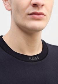 Tamsiai mėlynas ilgaamžis džemperis su grioveliu apykakle, kurio krašte išsiuvinėtas tekstūrinis pilkas "BOSS" logotipas. Lygi medžiaga, prigludusi silueto forma.