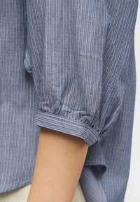 Manche en tissu rayé bleu-gris avec poignet froncé sur le bras d'une personne, montrant la texture et le motif à fines rayures verticales.