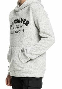 Persona che indossa un hoodie grigio chiaro di Quiksilver con tasca frontale e pantaloni cargo neri, mostrata dalla vita al petto, con la mano destra in tasca.