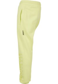Pantaloni de trening galbeni deschis, cu talie elastică, buzunare laterale și un logo imprimat vertical pe piciorul stâng. Textură moale și netedă.
