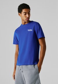 Mladík s krátkymi kučeravými vlasmi, ktorý má na sebe modré tričko Red Bull Racing a sivé tepláky, pozerá dole na svoju ľavú stranu.