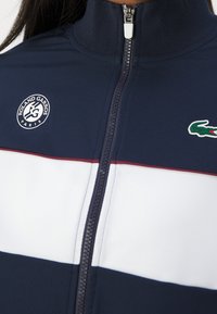 Marineblå zip-up jacka med vita och mörkblå horisontella ränder. Har en Lacoste krokodil-logotyp och Roland Garros-emblem på bröstet.