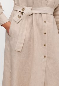 Abito in lino di un beige chiaro, caratterizzato da una cintura avvolgente, dettagli con bottoni, una tasca e una superficie testurizzata. I bottoni sono color ottone.