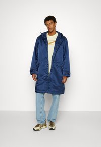 Parka impermeabile blu navy con cappuccio, bottoni frontali, due grandi tasche e una morbida fodera interna, indossata sopra una maglietta chiara e jeans.