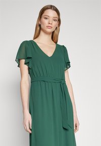 Rochie verde dintr-un material ușor, cu mâneci scurte cu volane, decolteu în V și o centură legată în talie, având o fustă fluidă.