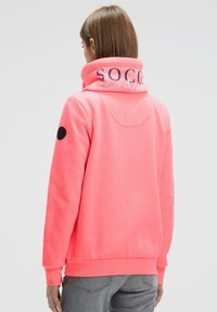 Neon pink hoodie with a high collar, featuring a soft texture, a logo in white print on the collar, and black patch accents on the sleeves.