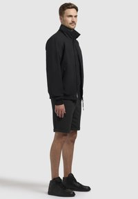 Chaqueta negra con cuello alto, bolsillos laterales y puños acanalados, combinada con pantalones cortos oscuros y zapatillas altas negras. Diseño simple.