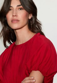 Haut rouge à manches bouffantes avec un décolleté rond. Tissu doux et texturé avec des détails froncés sur les manches. Grandes boucles d'oreilles en or.