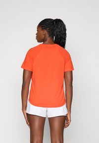 Kortermet treningstrøye i klar oransje stoff med rund hals og raglanermer, brukt med hvite shorts.