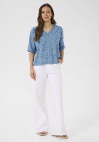 Svjetloplava bluzica kratkih rukava s V-izrezom i zatvaranjem na dugmad, s blagim teksturiranim uzorkom, u kombinaciji s bijelim hlačama širokih nogavica.