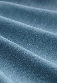 Tela de denim en azul claro con una textura suave, mostrando sutiles líneas diagonales y pequeñas motas, doblada en suaves ondas.