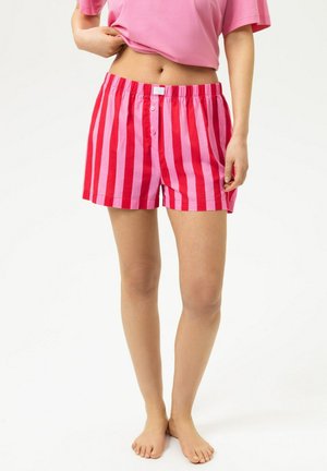 Personne portant un short rayé vertical rose et rouge et un haut court rose, debout pieds nus sur un fond blanc.