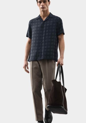 Man met een donker, patroonrijke, kort mouwen overhemd met knopen en lichtbruine broek, die een grote donkere draagtas in zijn rechterhand vasthoudt.