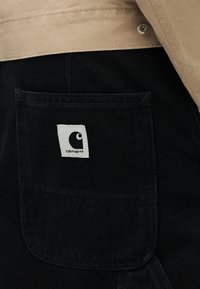 Κοντινή λήψη της τσέπης μαύρου τζιν παντελονιού με ένα λευκό έμβλημα λογότυπου Carhartt και μπεζ τελείωμα μπουφάν από πάνω.