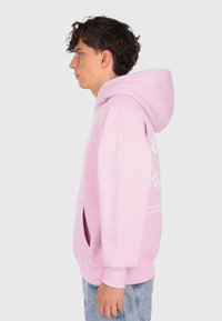 Giovane in felpa rosa con testo bianco sulle maniche e sul retro, in piedi di lato contro uno sfondo chiaro e uniforme.