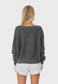 Blonde woman wearing a loose, dark gray knitted sweater and light gray shorts, standing facing away against a plain background.