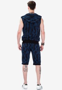 Blaues Camouflage-Ärmelloses Hoodie mit schwarzen Akzenten, kombiniert mit passenden Shorts mit umgeschlagenen Säumen. Getragen mit weißen Sneakers.