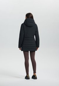 Zwarte pufferjack met een capuchon, voorzien van een gestructureerd, quilted ontwerp en een aantrekbare taille, gedragen over bruine leggings en zwarte sneakers.