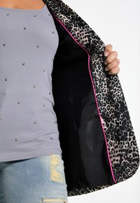 Chaqueta de estampado de leopardo con forro negro, que presenta un ribete de color rosa brillante, llevada sobre una blusa gris de canalé con adornos plateados.