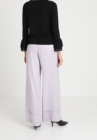 Zwarte trui met ruches aan de mouwen en decoratieve accenten, gecombineerd met lichtlila wijde broek en zwart met hoge hakken.